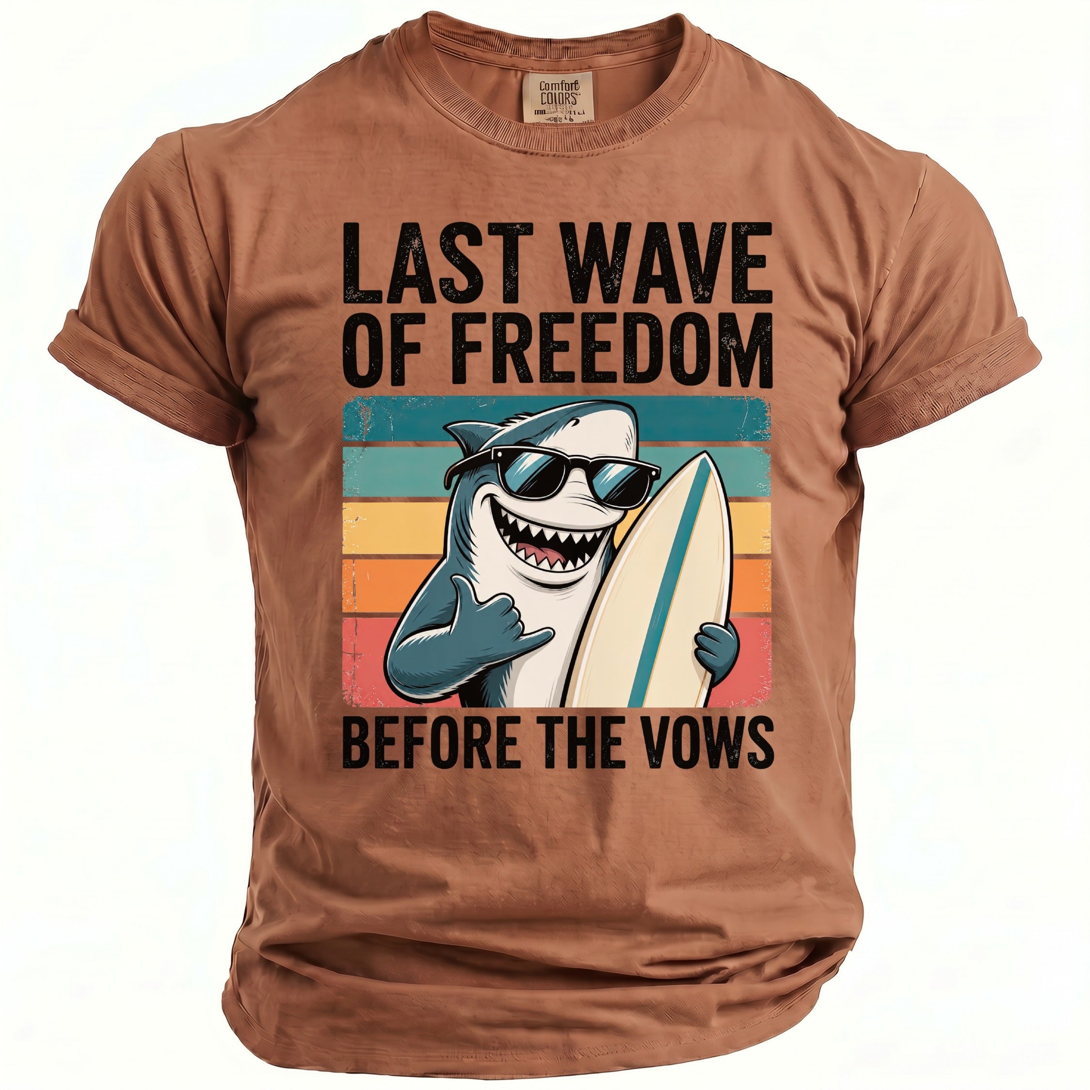 Brown t-shirt with graphic of a shark wearing sunglasses holding a surfboard. Text reads "LAST WAVE OF FREEDOM BEFORE THE VOWS".