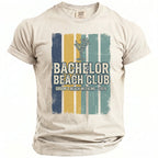 Cream t-shirt with "Bachelor Beach Club Groom's Beach Weekend 2026" text and graphics.