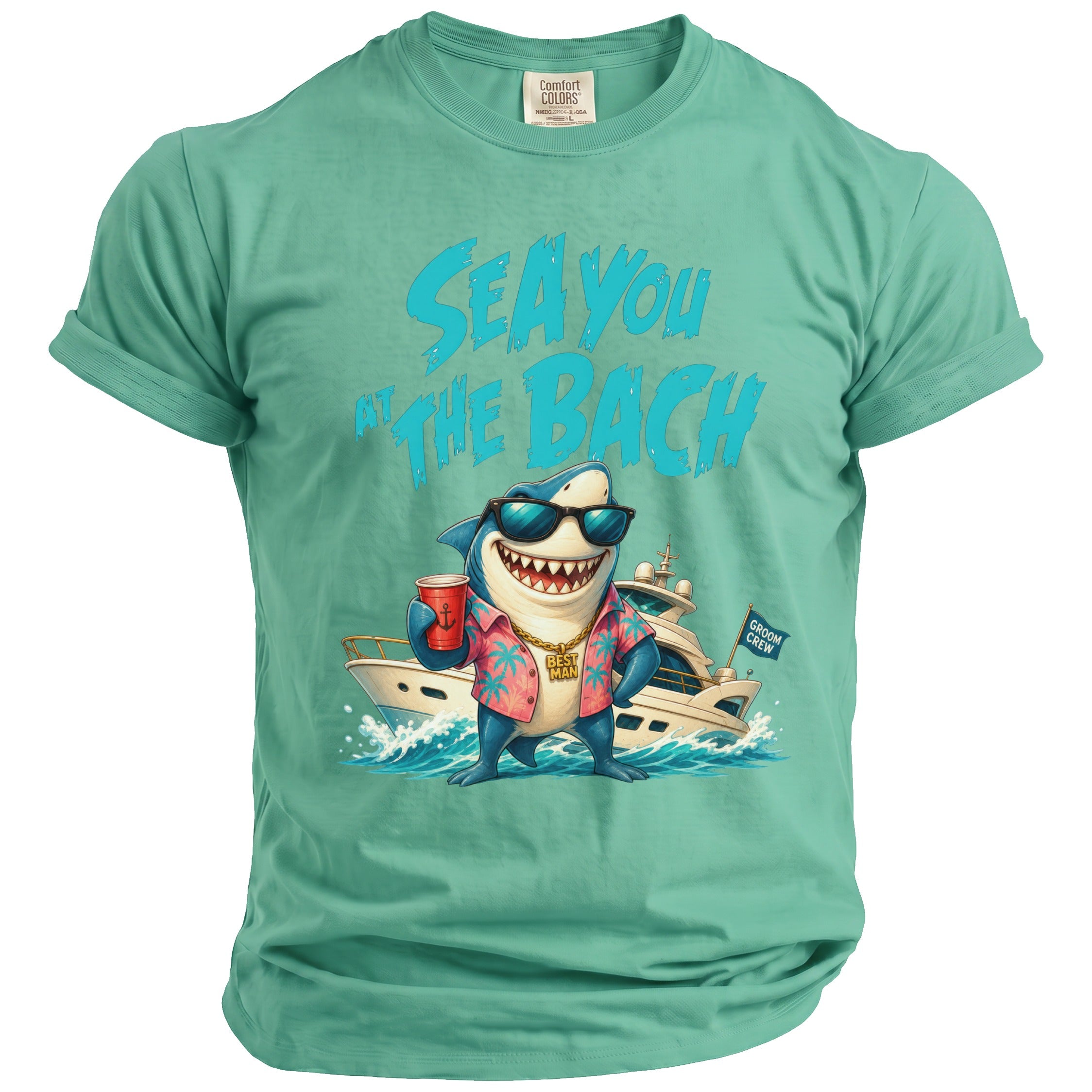 A green short-sleeve t-shirt features a graphic of a smiling shark wearing sunglasses and a pink aloha shirt, standing in front of a yacht with text reading Sea You At The Bach above.